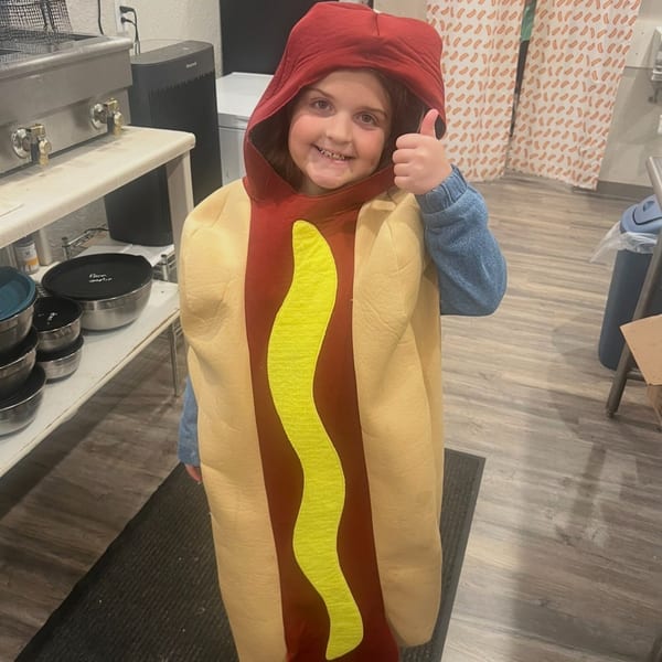 A child in a hot dog costume with mustard gives a thumbs up in a restaurant kitchen.