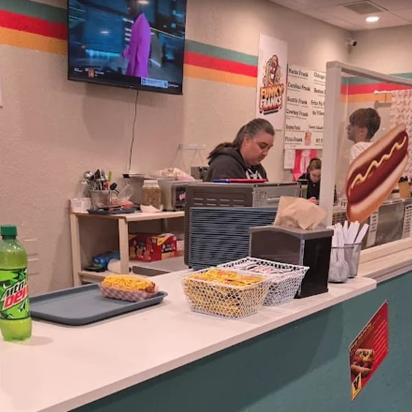 A Funky Franks employee works behind the counter, with a hot dog and Mountain Dew on the counter.
