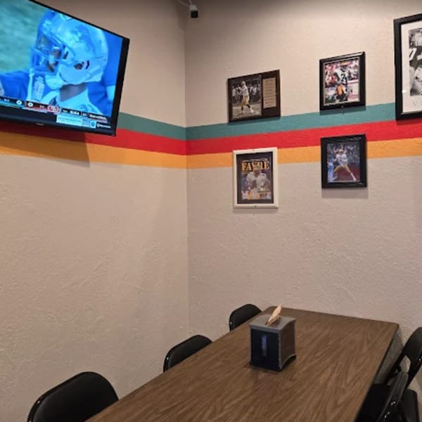 Corner of a restaurant dining area with a TV, sports photos, and a wooden table with chairs.