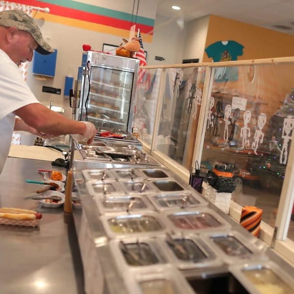A Funky Franks employee prepares hot dogs at a counter with various toppings and a hot dog warmer.