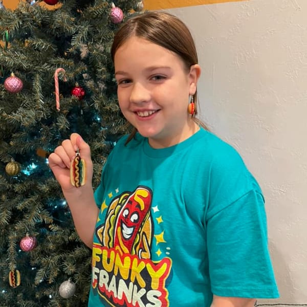 Smiling girl in a Funky Franks shirt holding a hot dog ornament next to a Christmas tree.
